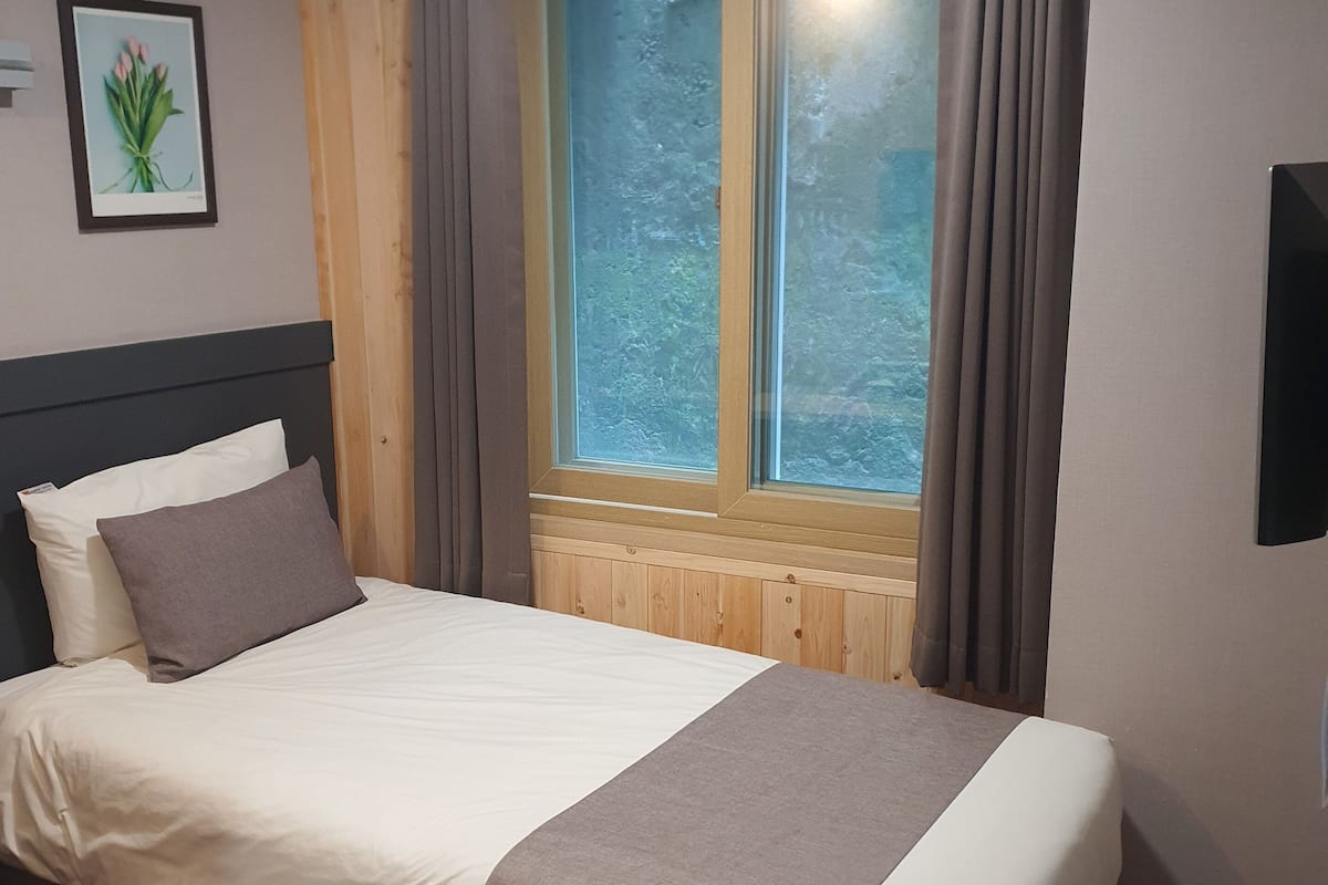 A neatly made bed with a white and brown bedspread, a headboard, and a bedside table with a lamp. There is a window with curtains, a framed picture on the wall, and a flat-screen TV mounted on the wall.