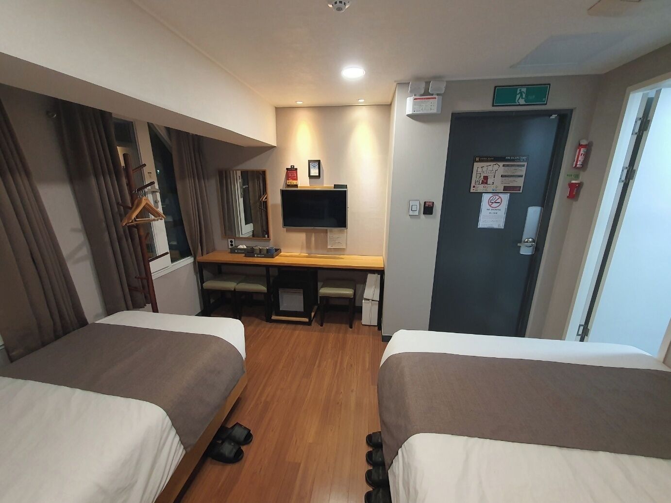 A hotel room with two beds, a desk with a computer, a mirror, and a door with emergency exit signs.