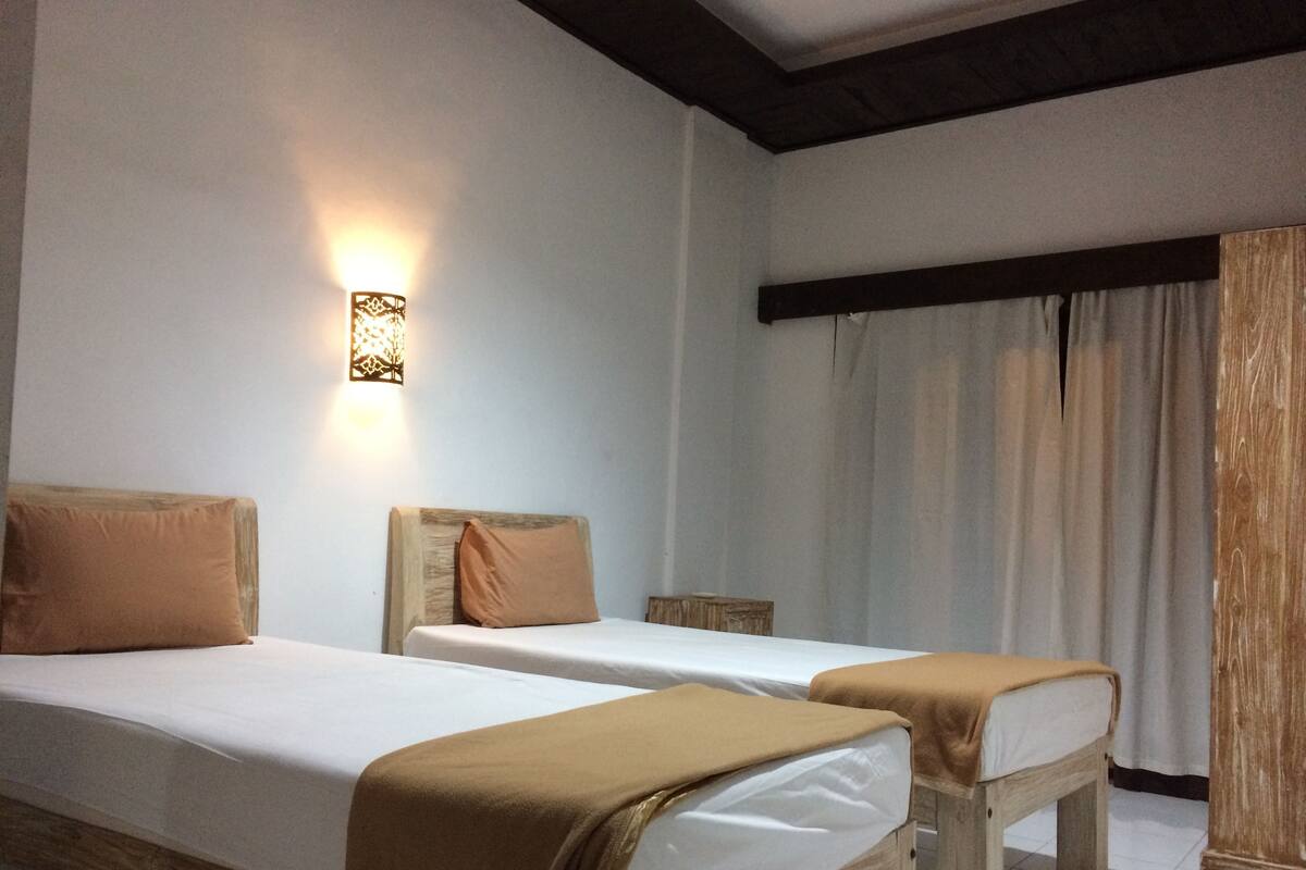A hotel room with two single beds, a wooden headboard, a wall-mounted light fixture, and a window with curtains.