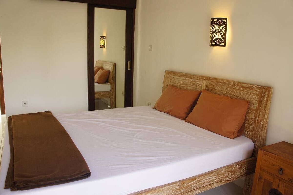 A neatly made bed with a brown blanket, a wooden headboard, and two orange pillows. There is a wooden nightstand to the right and a wall-mounted light fixture above the bed.