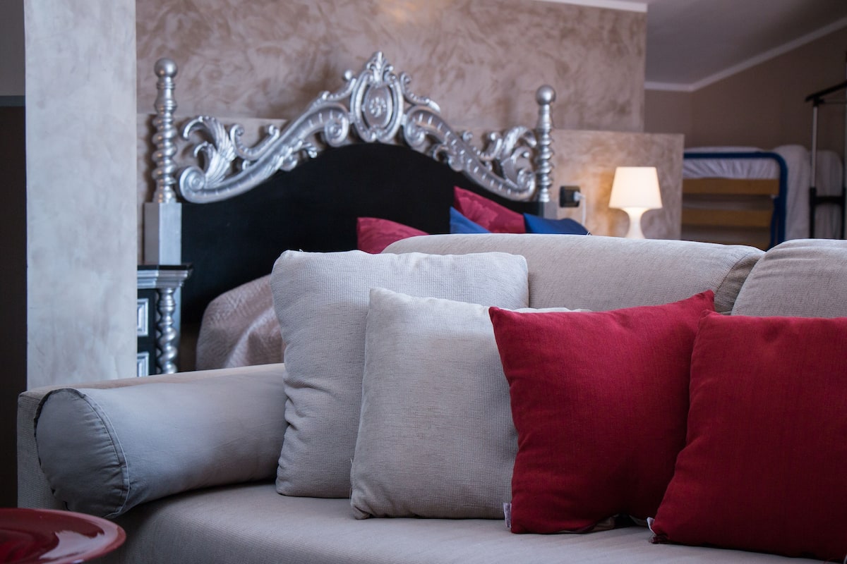 A hotel room with a bed featuring an ornate headboard, a sofa with multiple cushions, and a bedside table with a lamp.