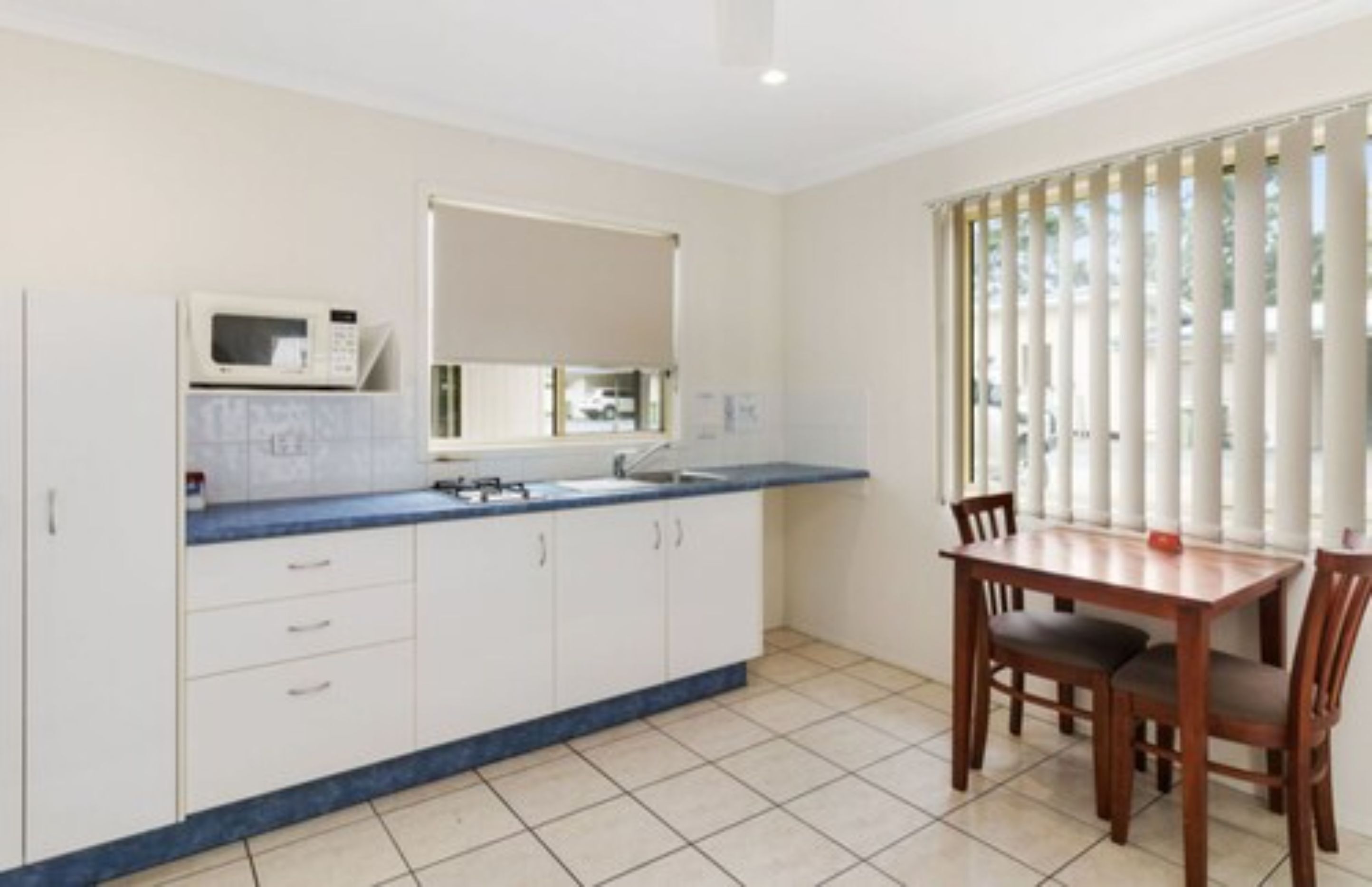 A kitchen with a microwave, sink, and cabinets. There is a dining table with chairs and a window with blinds.