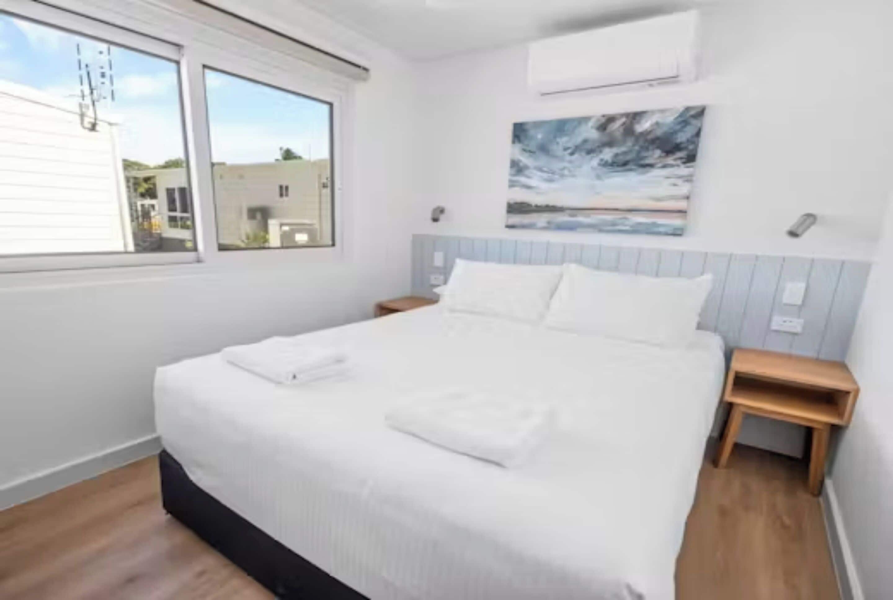 A bedroom with a bed, a nightstand, a window, and a painting on the wall.