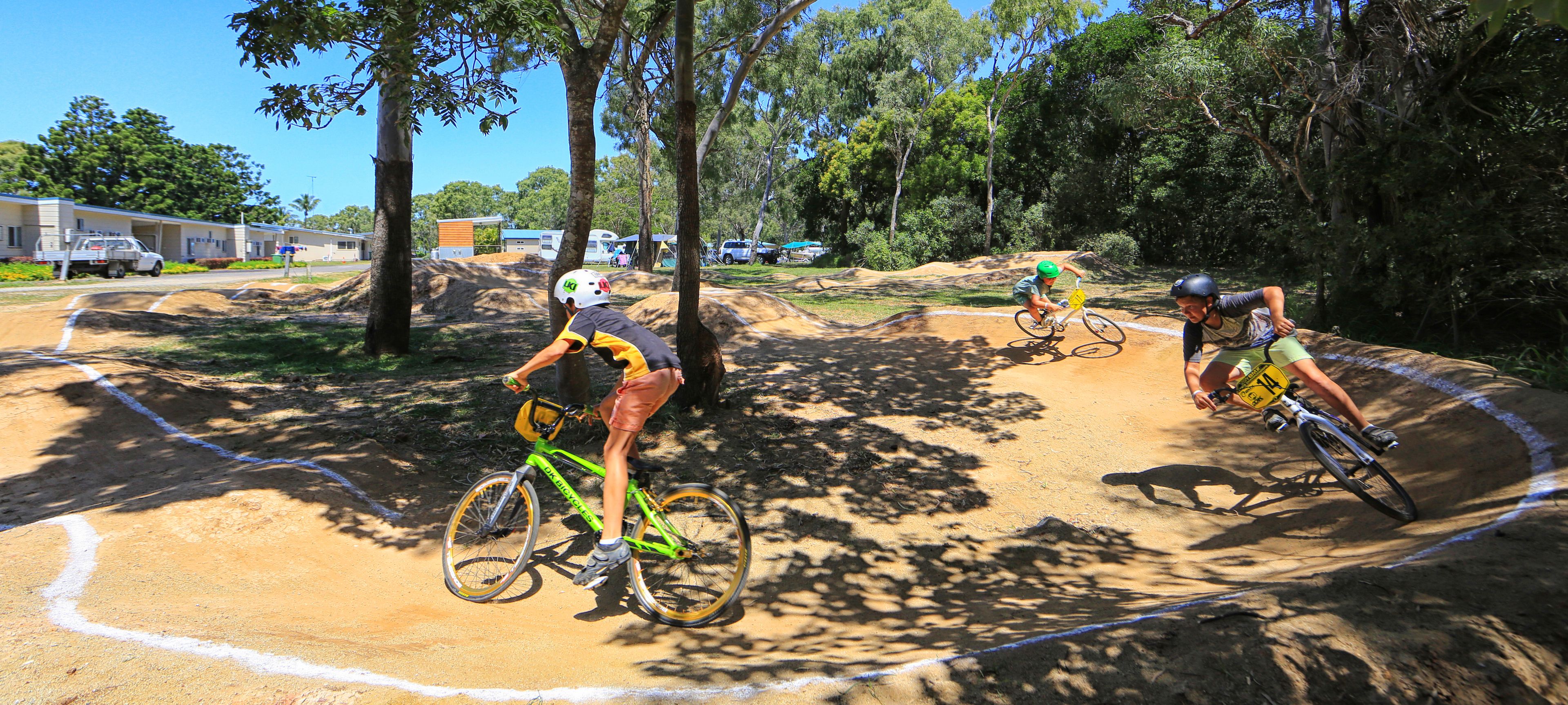 A dirt bike track with riders, surrounded by trees and a building in the background.