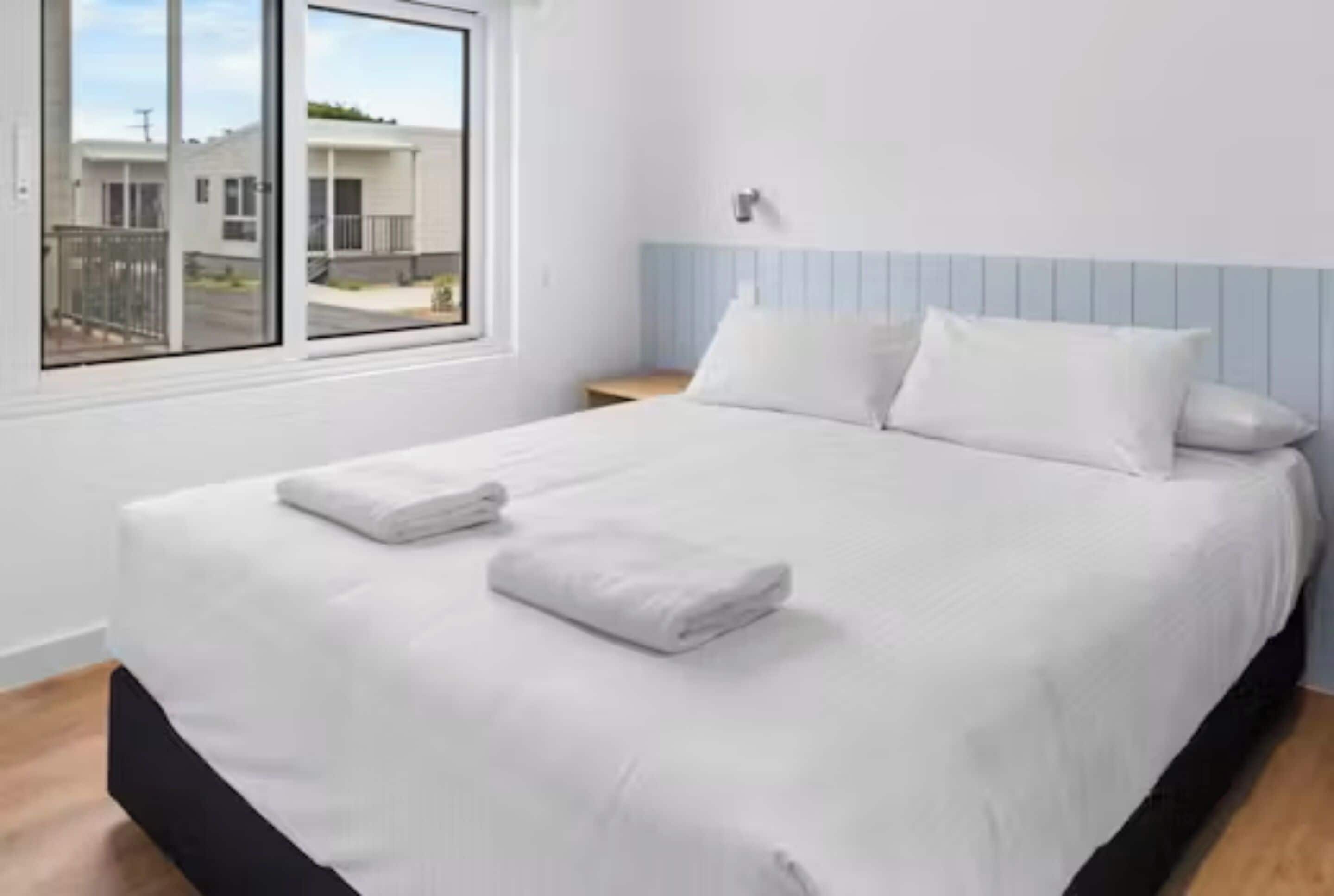 Una cama con sábanas blancas y dos almohadas, una ventana con vista a edificios y una pared de color azul claro.