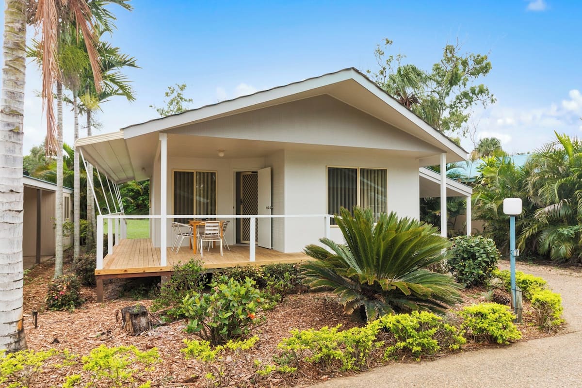 A single-story house with a covered porch, a small deck, and a garden with various plants and a palm tree.