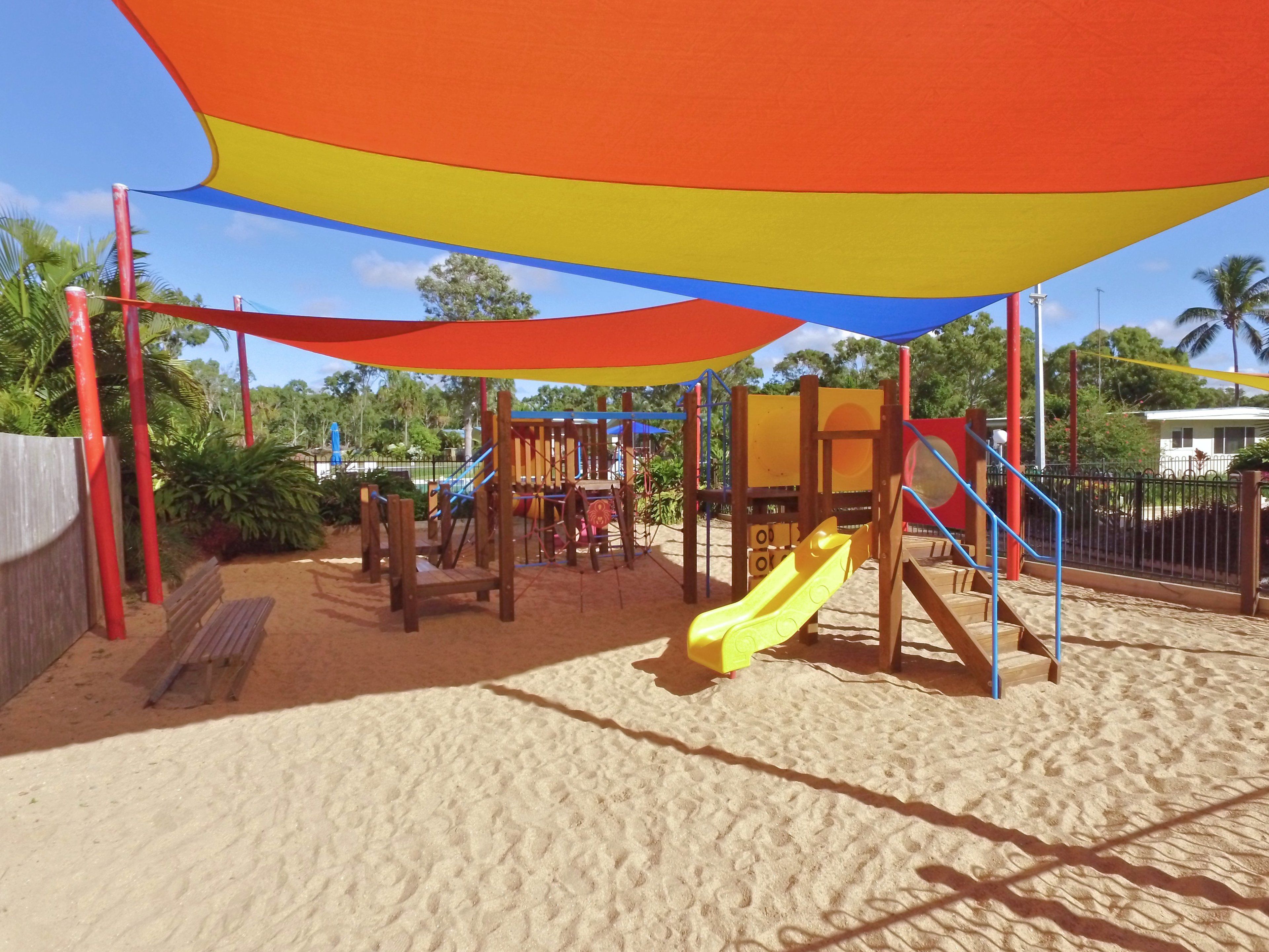 A playground with a yellow slide and climbing structures, set on sandy ground with a colorful canopy overhead.
