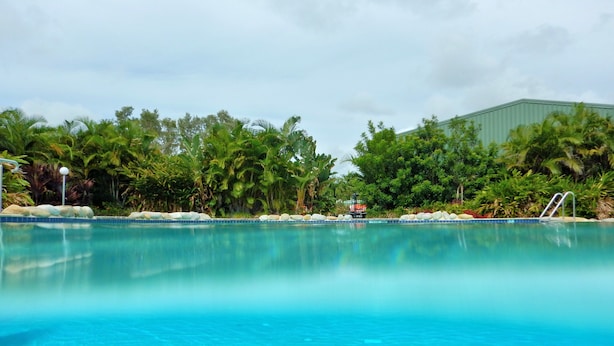 A clear blue pool surrounded by lush greenery and a building in the background. - Discovery Parks - Coolwaters Yeppoon (Kinka Beach)
