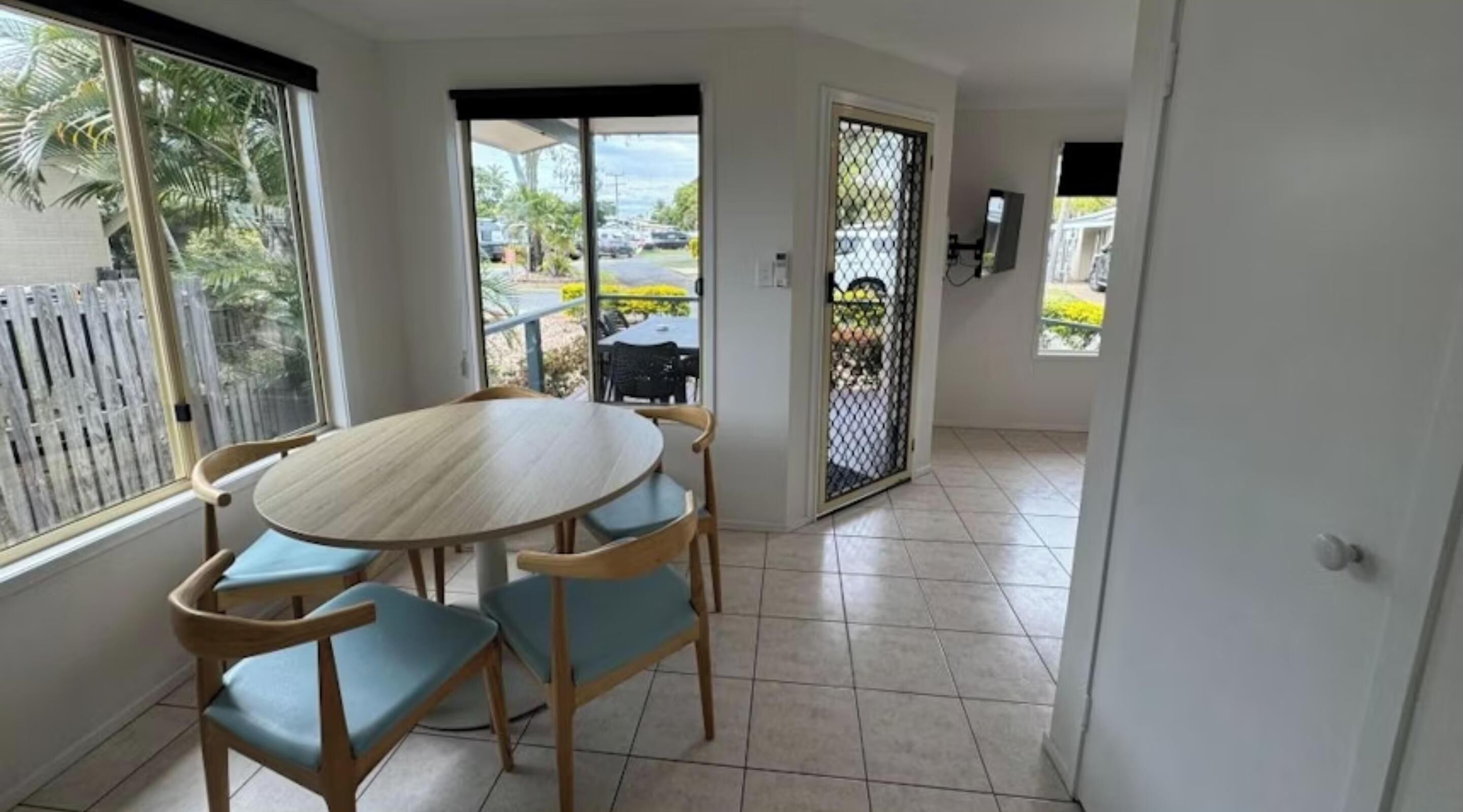 Una habitación con una mesa redonda de madera y cuatro sillas, un ventanal corredero que da al exterior, y piso de baldosas.