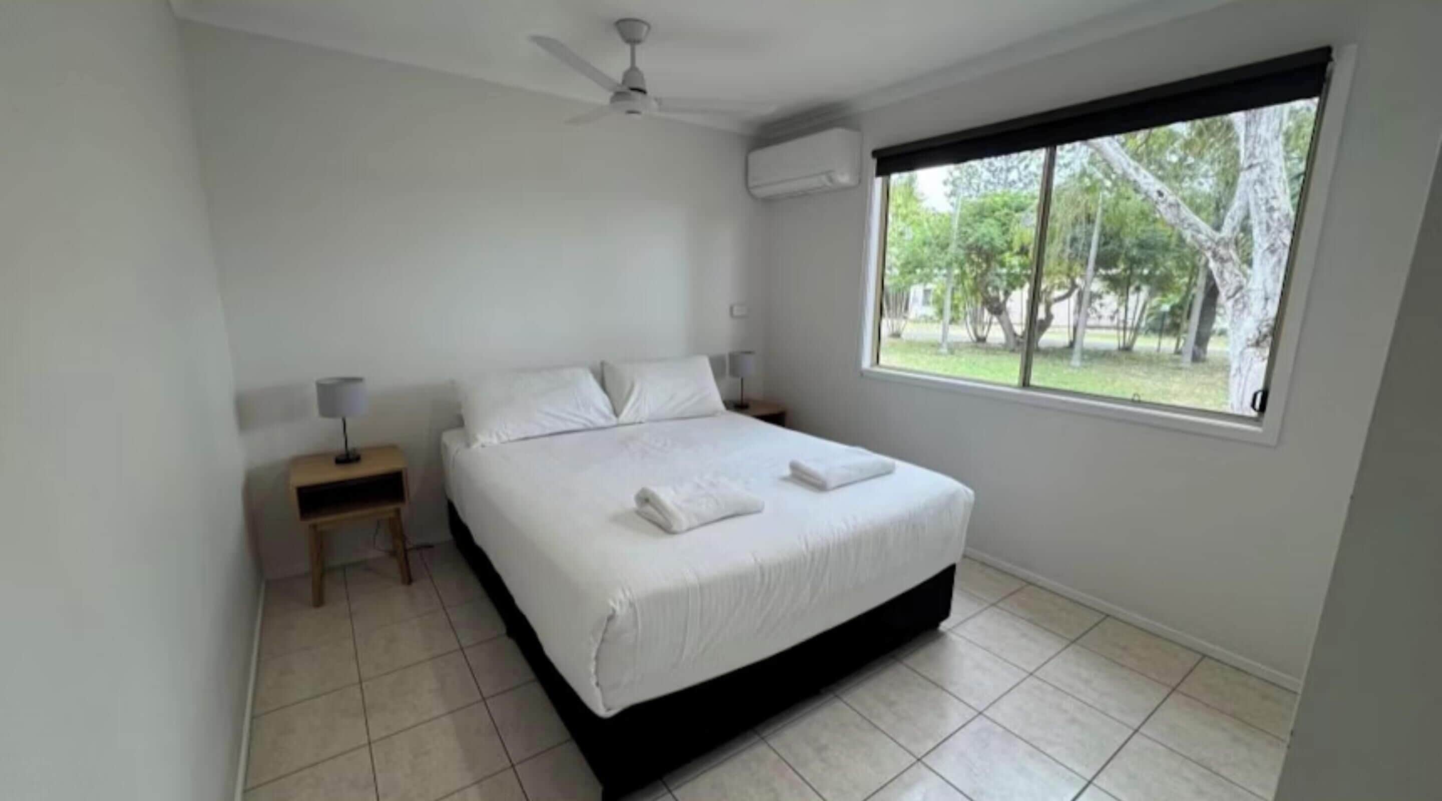A bedroom with a bed, a nightstand, a lamp, a ceiling fan, an air conditioning unit, and a window with a view of trees.