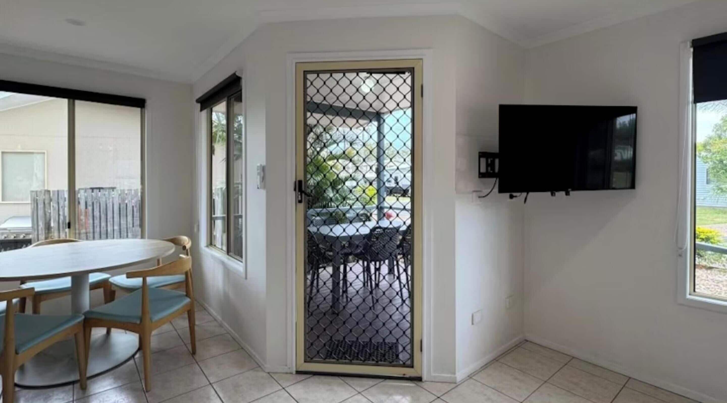 Quarto com mesa de jantar e cadeiras, televisão fixada na parede e porta que dá para o exterior.