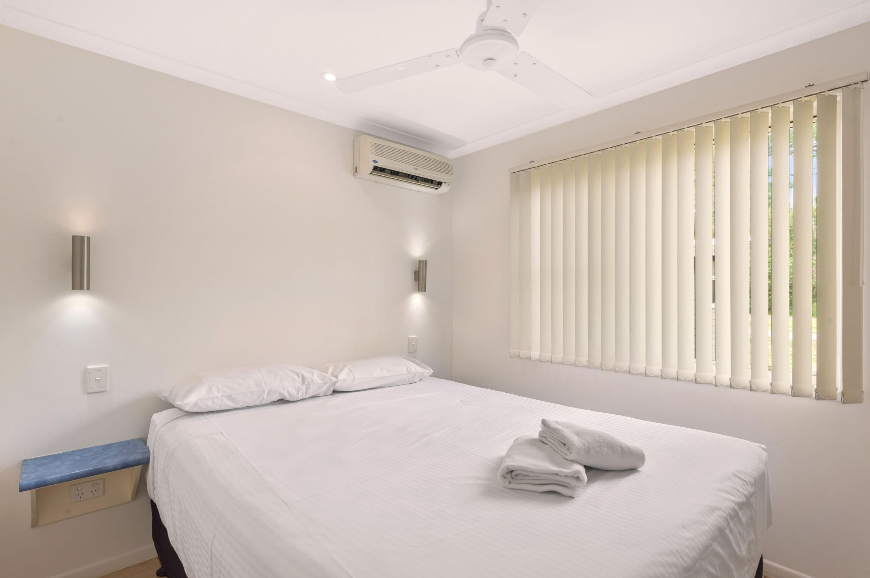 A hotel room with a bed, a ceiling fan, an air conditioner, a window with blinds, and a bedside table with a lamp.
