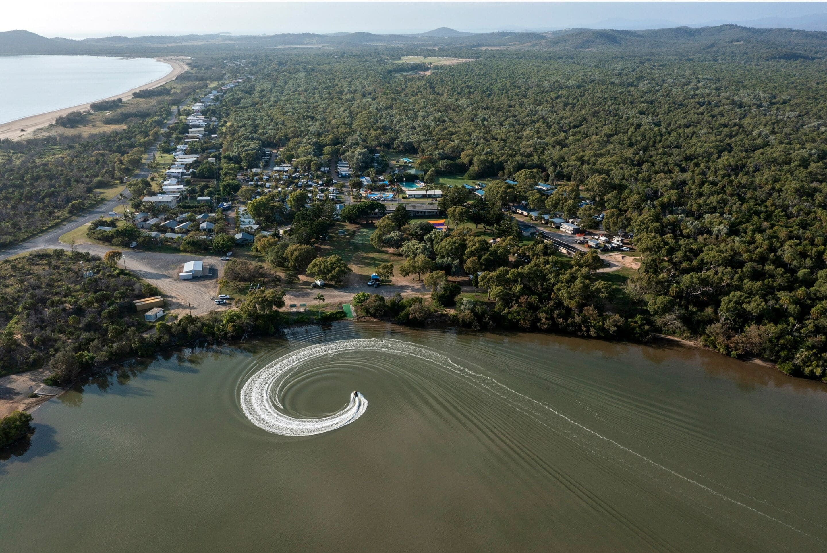 Vista aérea de una zona costera con una hilera de casas a lo largo de la orilla, cielo despejado y un barco dejando estela en el agua.