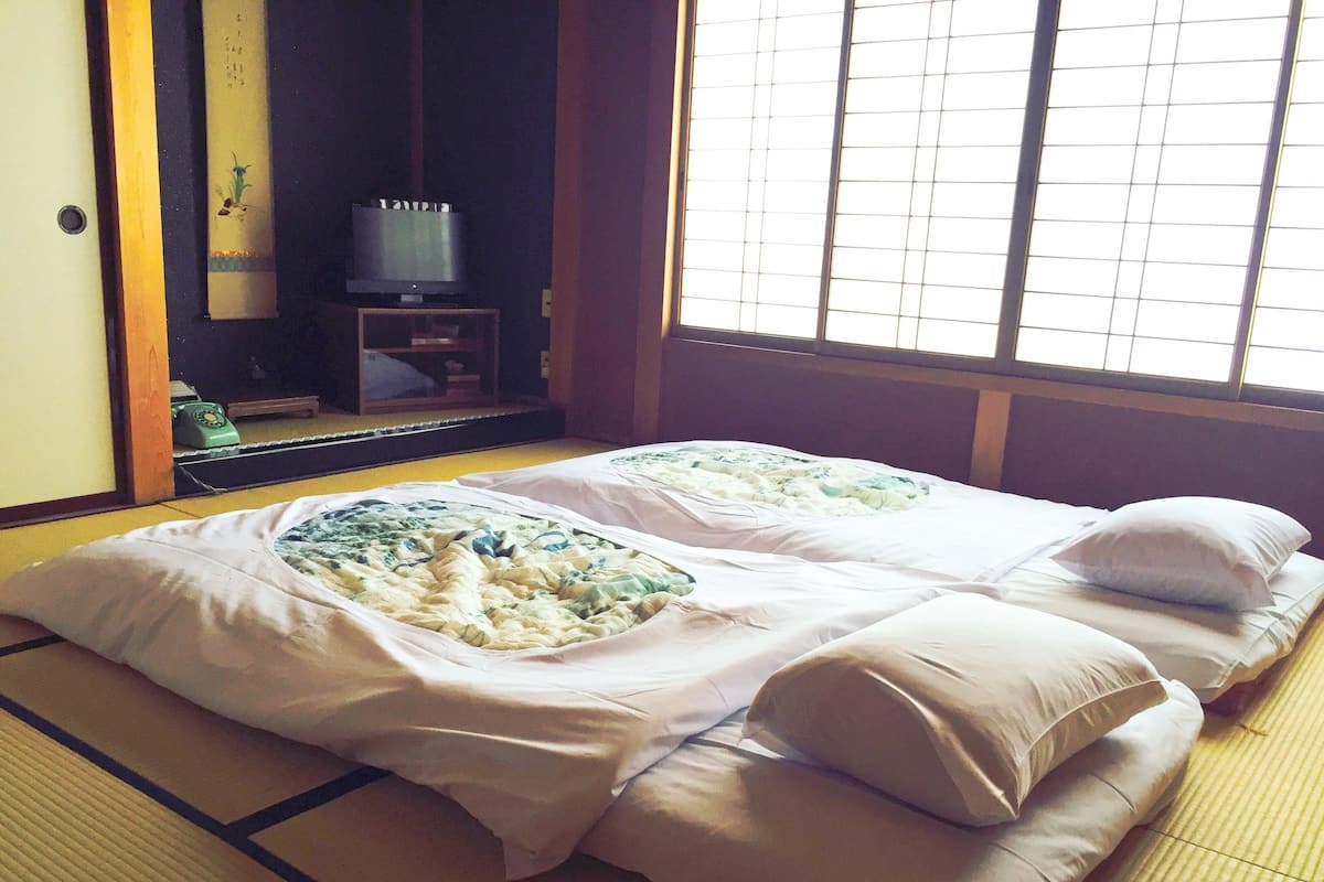 Een traditionele Japanse kamer met tatami vloeren, twee tatami matten, een futonbed met een wit en groen bloemenbedekking en een televisie op een houten standaard.
