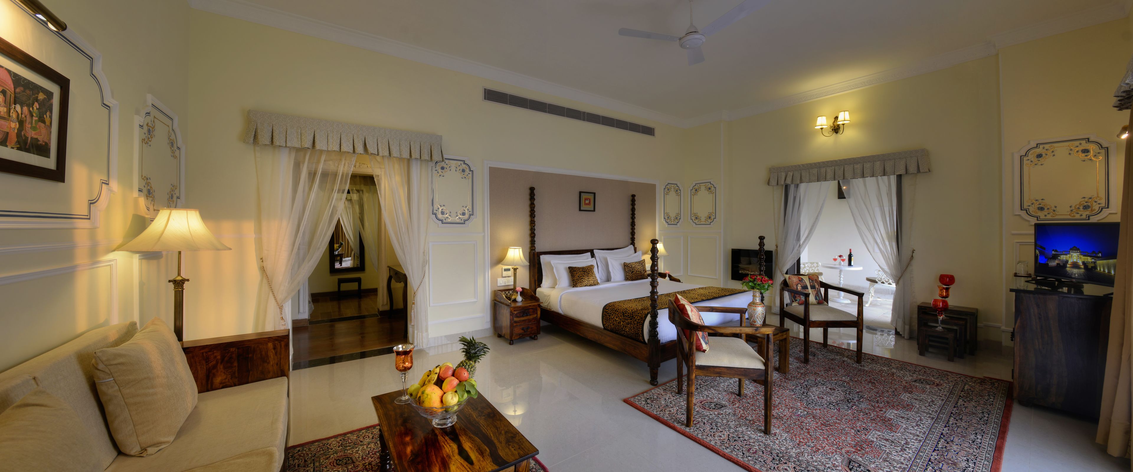 Quarto de hotel com uma cama, área de estar com sofá e poltrona, mesa de jantar com uma tigela de frutas e uma televisão.