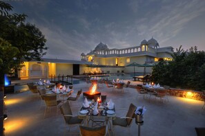 An outdoor dining area with tables and chairs, a fire pit, and a pool in the background. - The Tigress Resort & Spa, Ranthambore (Sawai Madhopur)