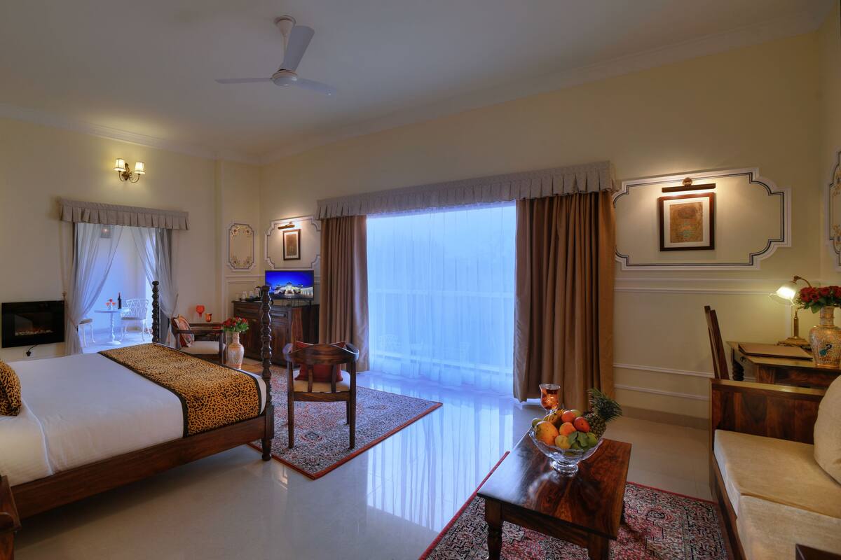 A hotel room with a large bed, a desk, a chair, a TV, and a balcony with curtains.