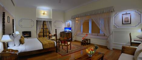 Une chambre d’hôtel élégamment aménagée, dotée d’un grand lit, d’un coin salon avec un canapé et des fauteuils, d’une petite table sur laquelle se trouve un panier de fruits, et d’un téléviseur.