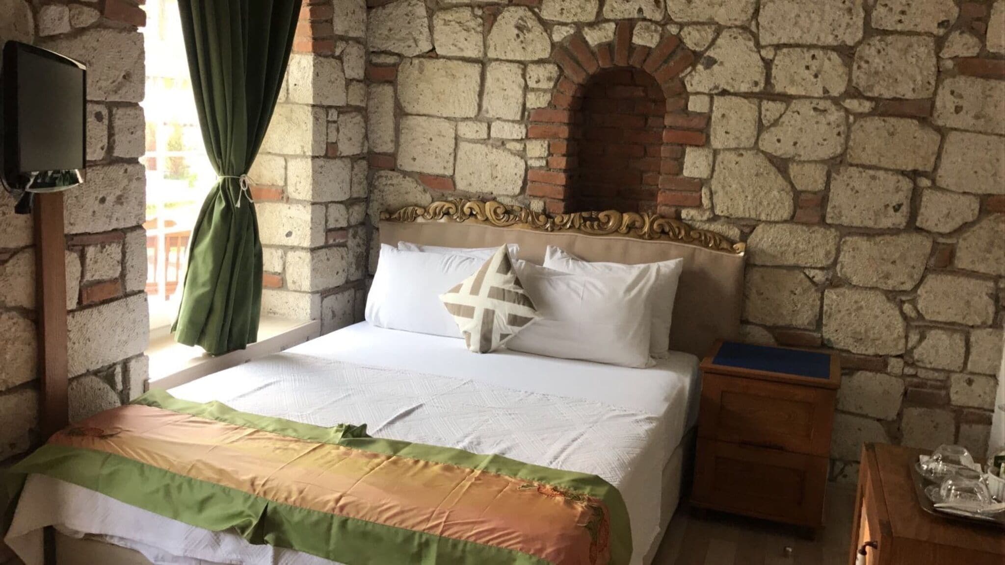 Un dormitorio con pared de piedra, una cama con ropa de cama blanca, una mesita de noche de madera y un televisor en la pared.
