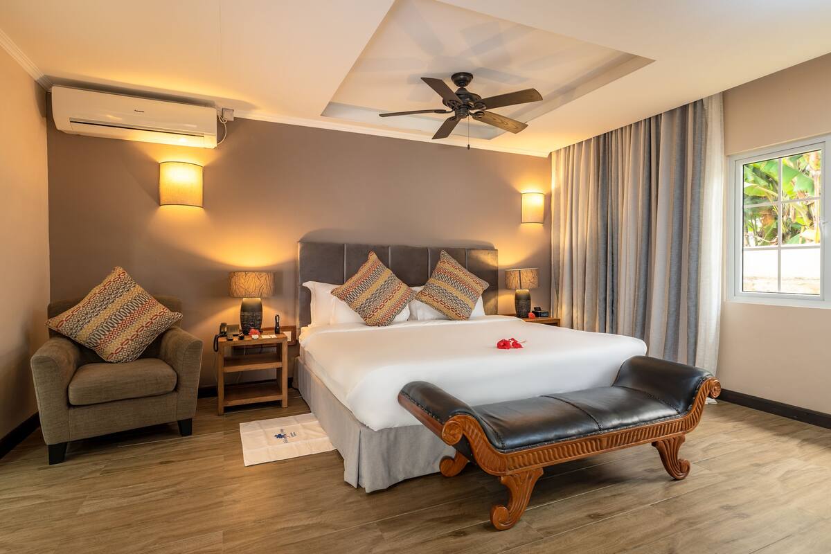 A bedroom with a large bed, a chair, a bedside table, a lamp, a ceiling fan, and a window with curtains.