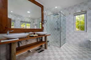 A modern bathroom with a wooden vanity, a large mirror, a glass shower enclosure, and a window with a view of greenery. - Felicie Cottage & Residence (Mahé Island)