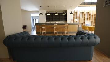 A modern living room with a dark blue sofa, wooden flooring, a kitchen area with a bar, and pendant lighting.