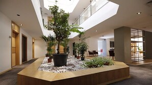 Un vestíbulo moderno con un mostrador de recepción de madera en el centro, rodeado de plantas en macetas y un camino de guijarros blancos.