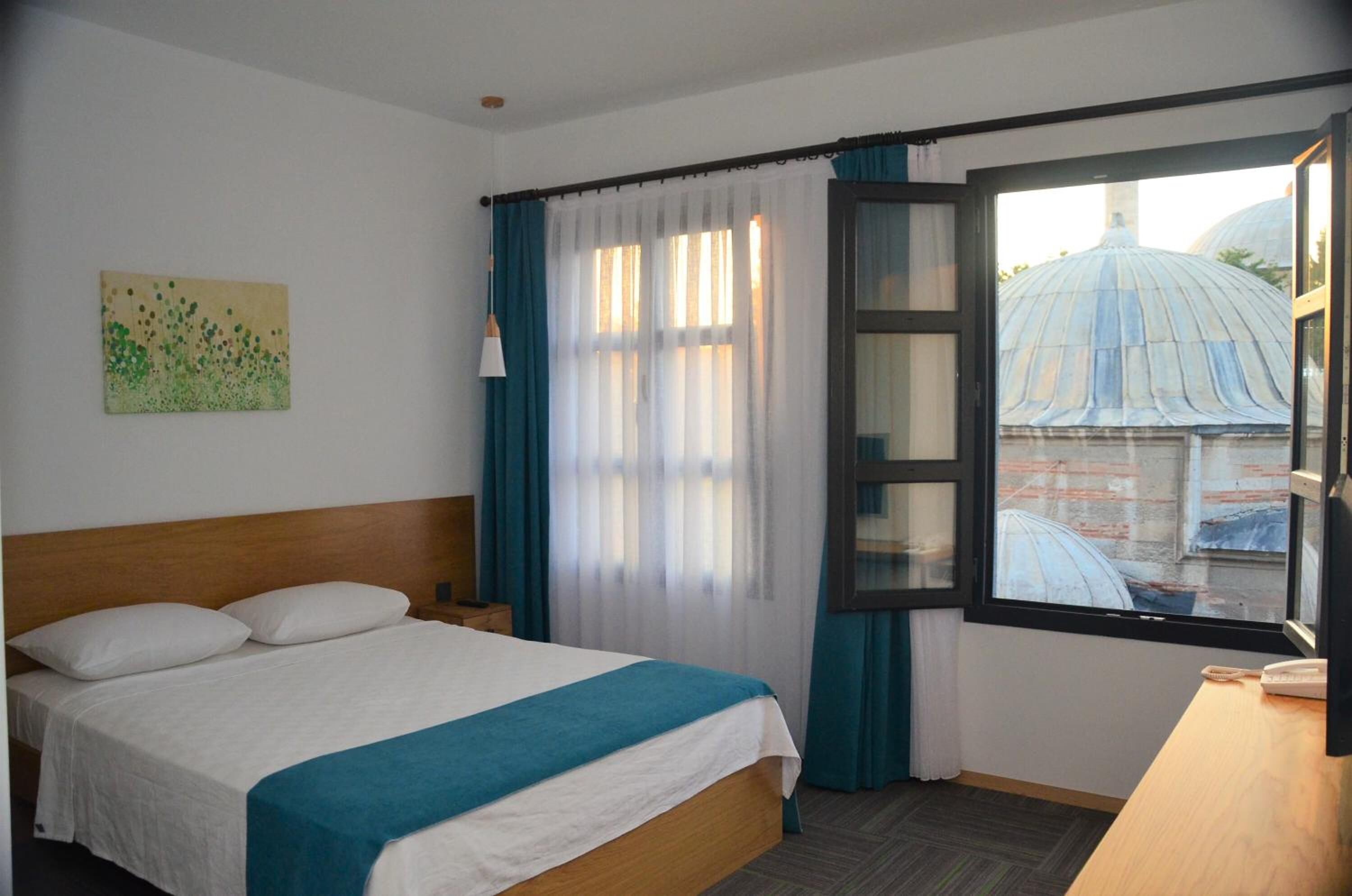 A hotel room with a bed, a window with curtains, a view of a dome structure, and a painting on the wall.