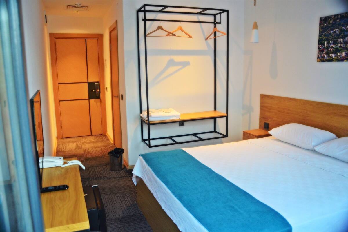 A hotel room with a bed, a bedside table, a hanging clothes rack, and a door.