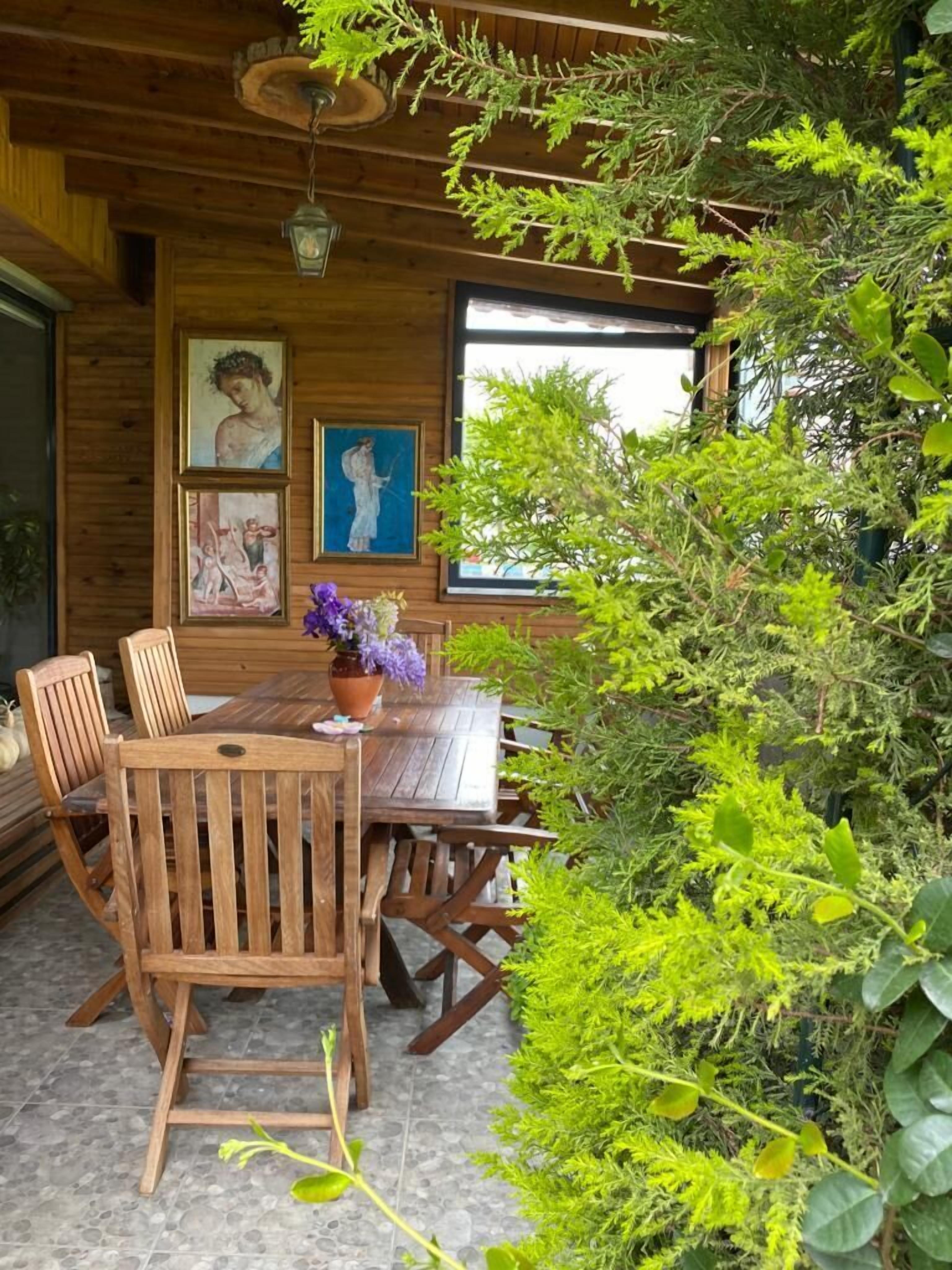 A wooden dining table with chairs, surrounded by greenery and framed artwork on the wall.