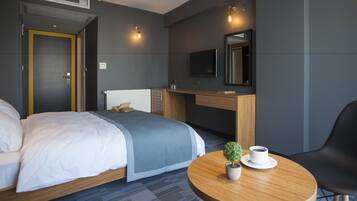A modern hotel room with a bed, bedside table, chair, and a small round table with a plant and cups.