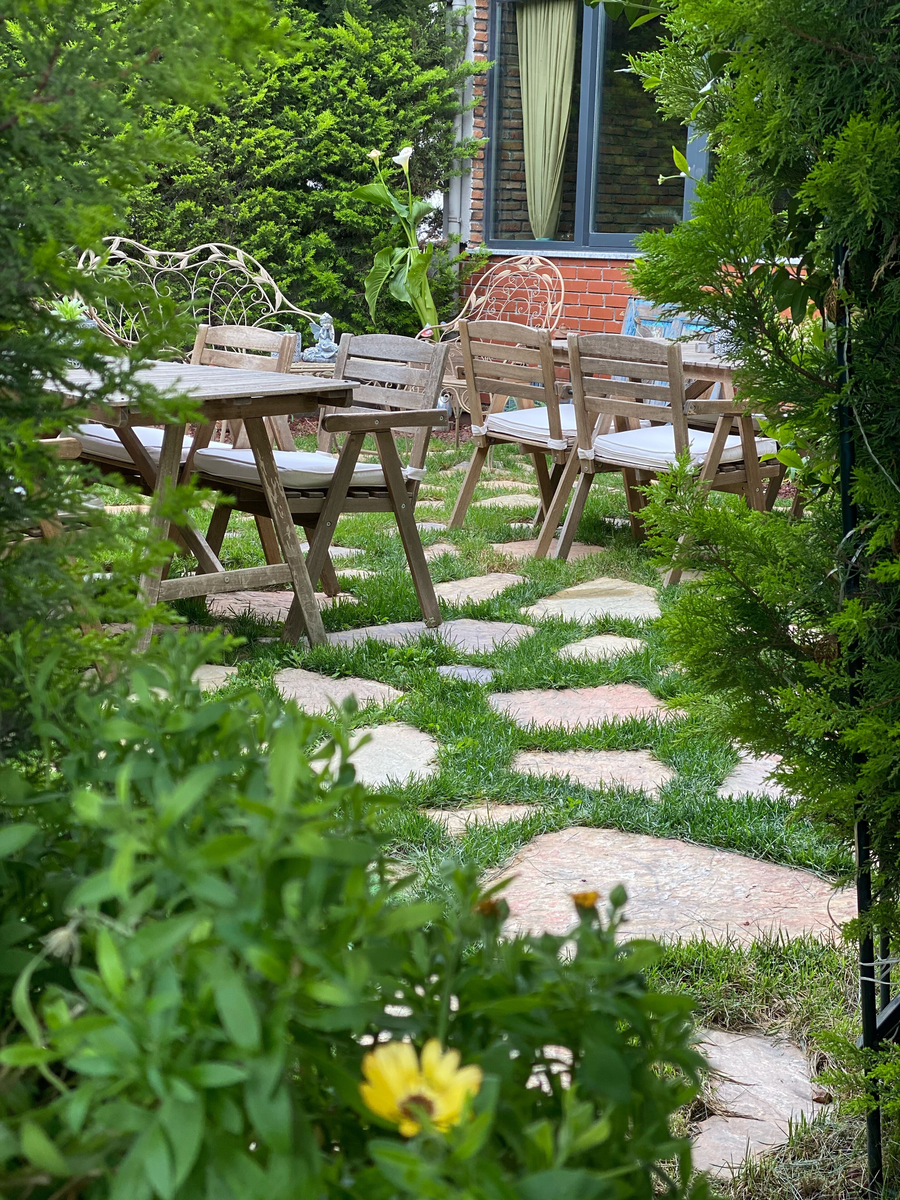 An outdoor seating area with wooden tables and chairs, surrounded by greenery and a stone pathway.