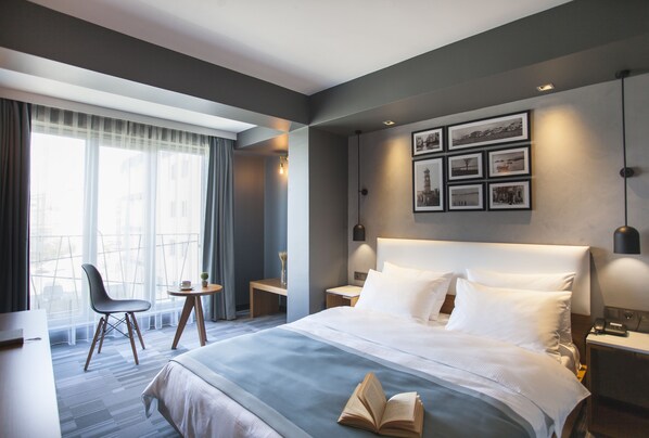 A modern hotel room with a large bed, a desk, a chair, and a view of the city through the window. - Hotel Cura (Çanakkale)