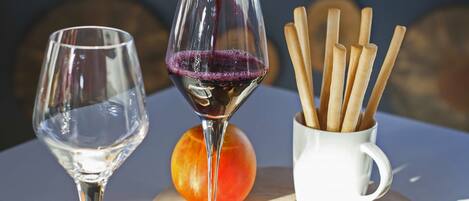 A table with two wine glasses, one filled with red wine and the other empty. There are fruits like apples and grapes, and a white mug with wooden sticks.