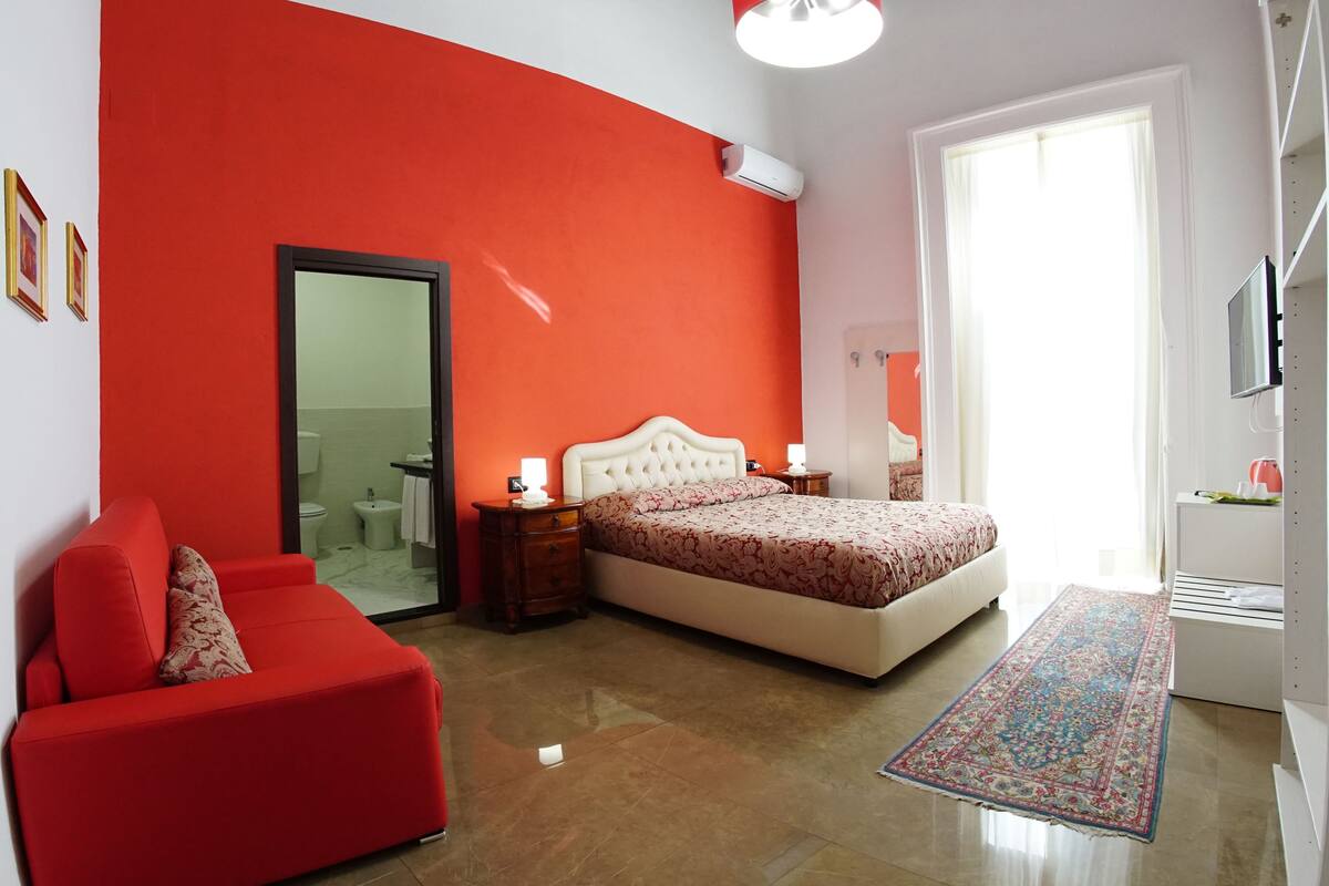 A bedroom with a red wall, a bed with a floral bedspread, a red sofa, a nightstand with a lamp, and a bathroom visible through a glass door.