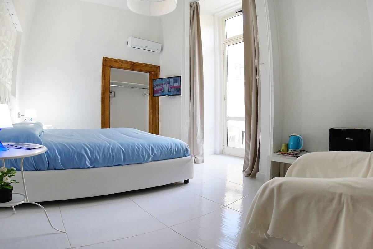 A hotel room with a bed, a desk, a chair, a potted plant, and a window with curtains.