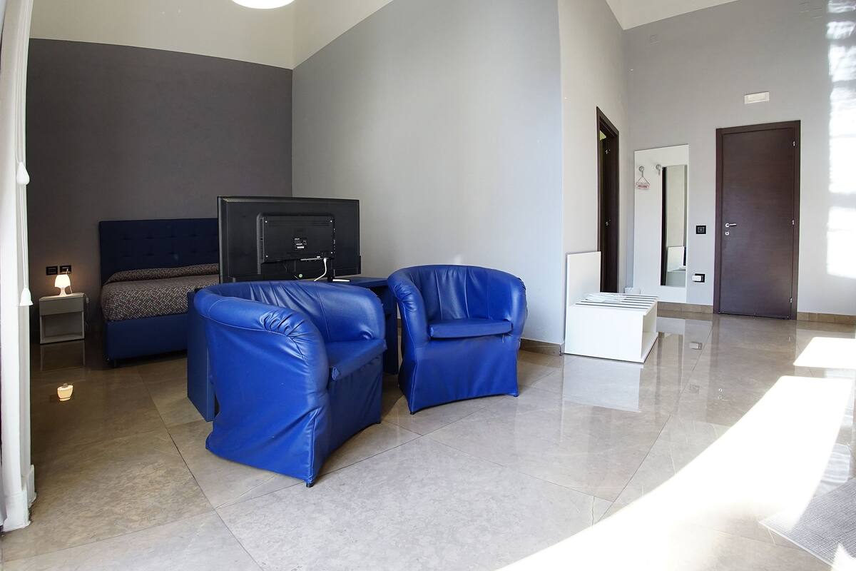 A modern hotel room with a bed, two blue armchairs, a flat-screen TV, and a small table.