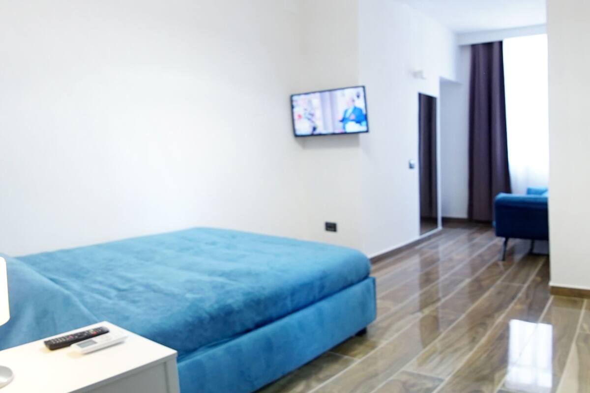 A modern hotel room with a blue bed, a white bedside table, a flat-screen TV mounted on the wall, and a wooden floor.