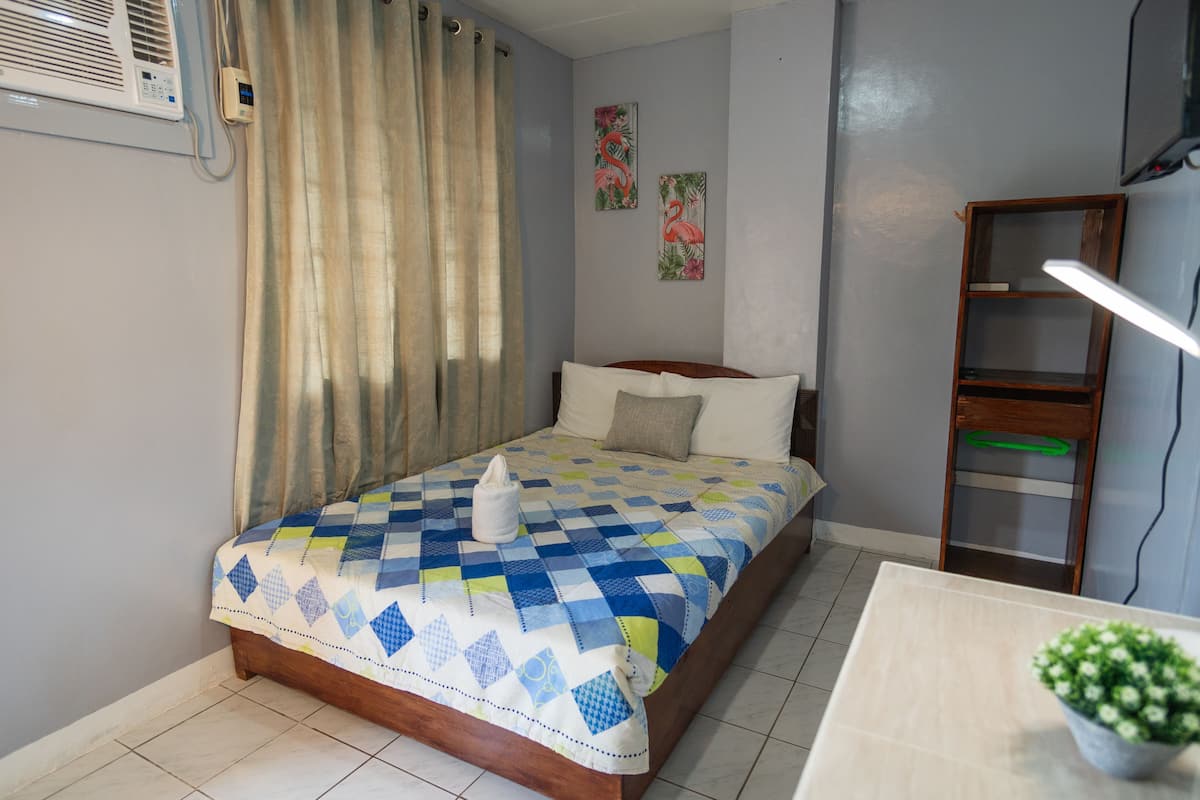 A bedroom with a bed, a bedside table, a lamp, a wall-mounted air conditioner, and a curtain.