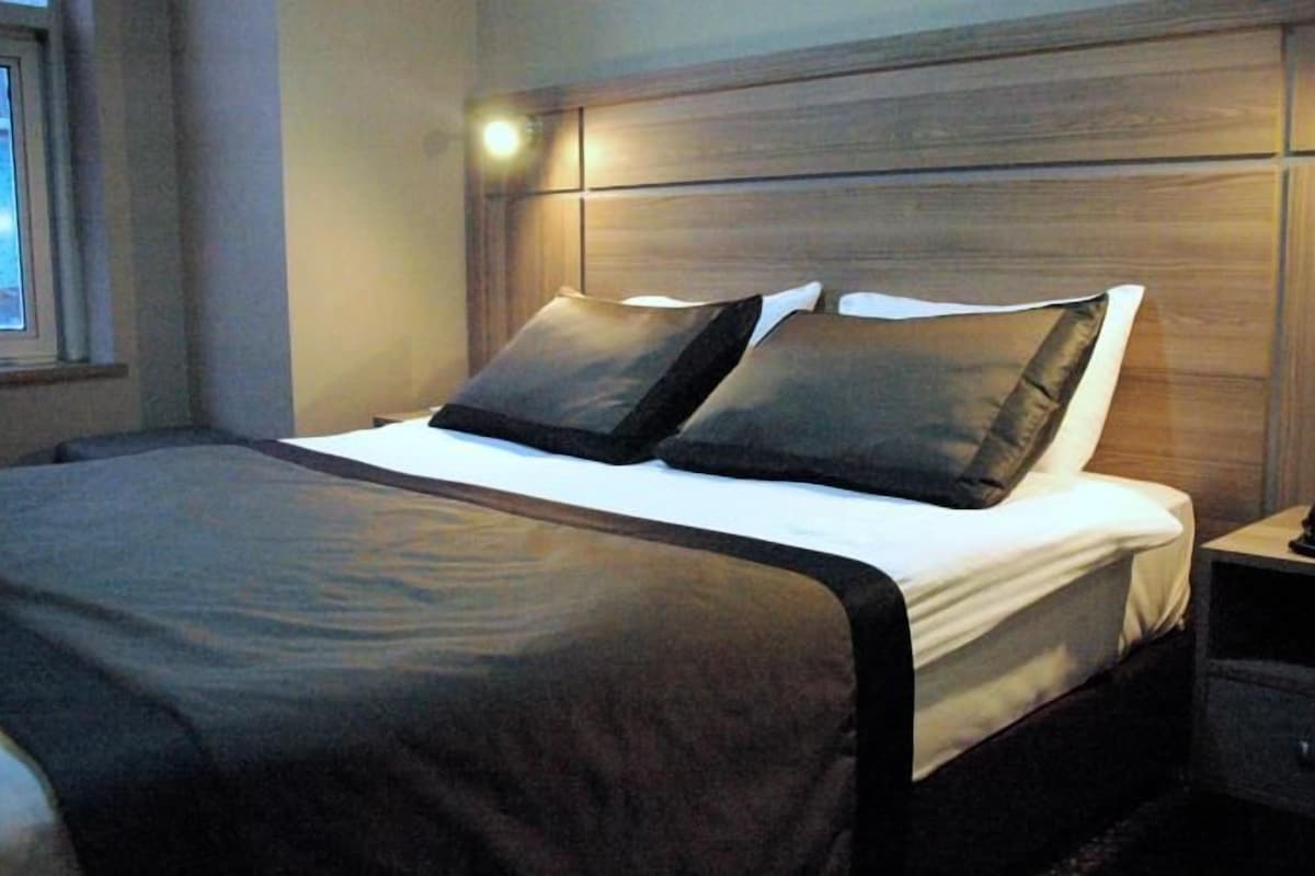 Een net opgemaakt bed met een donker hoofdbord, een nachtkastje met een telefoon, en een raam aan de linkerkant.