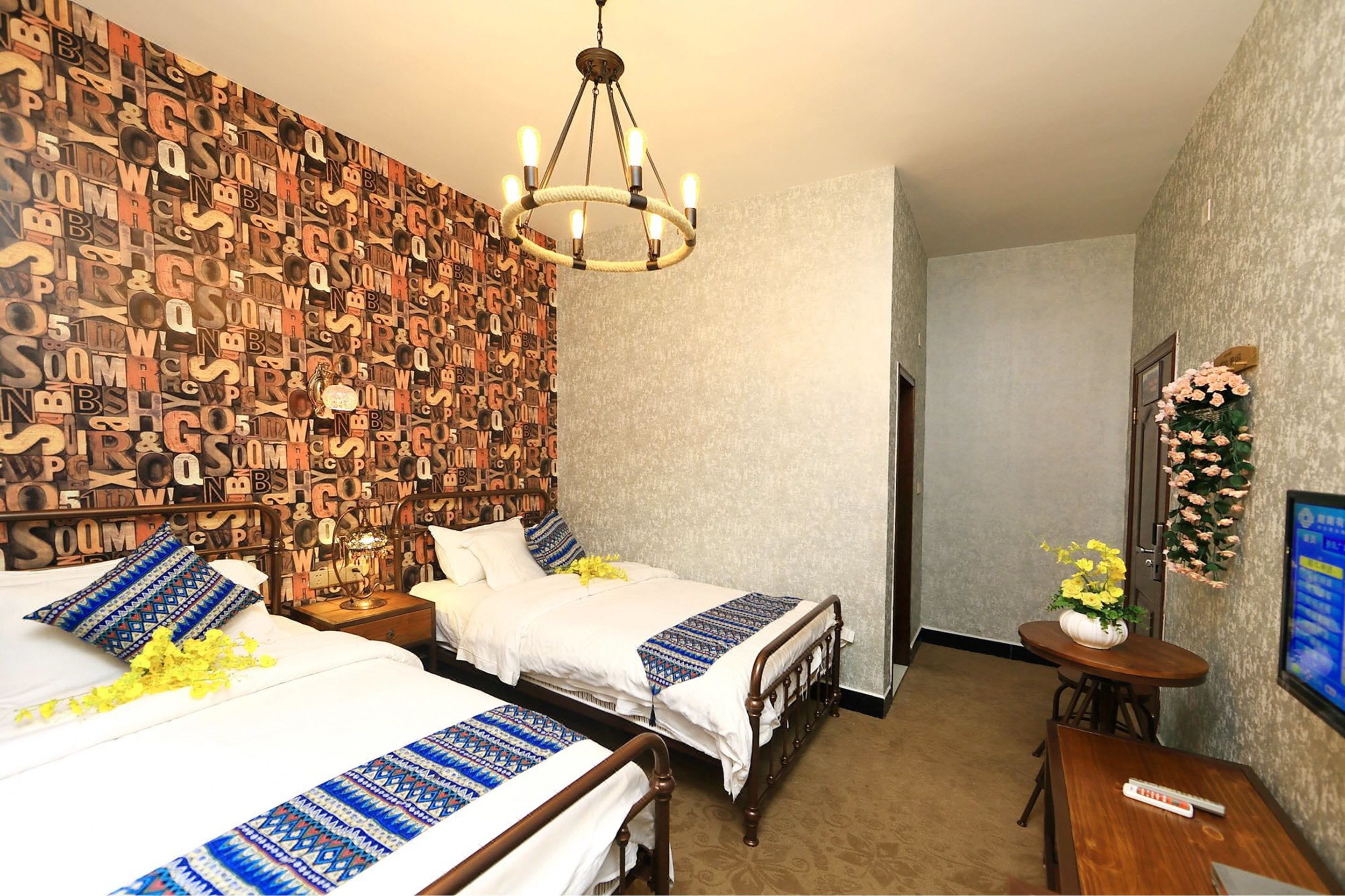 A hotel room with two beds, a chandelier, a small table with a vase of flowers, and a wall with a pattern of letters.