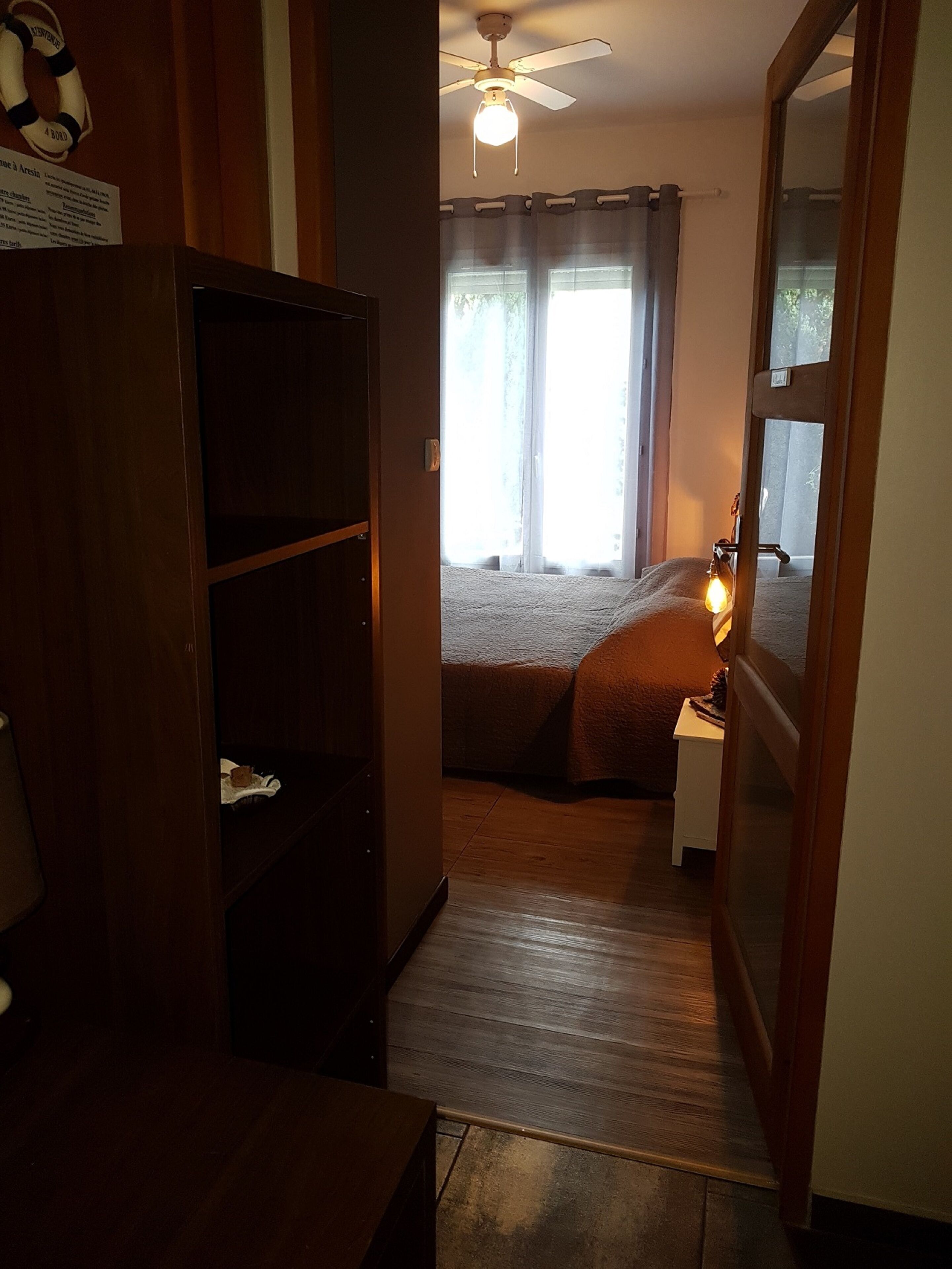 A narrow hallway leading to a bedroom with a bed, a nightstand, and a ceiling fan.
