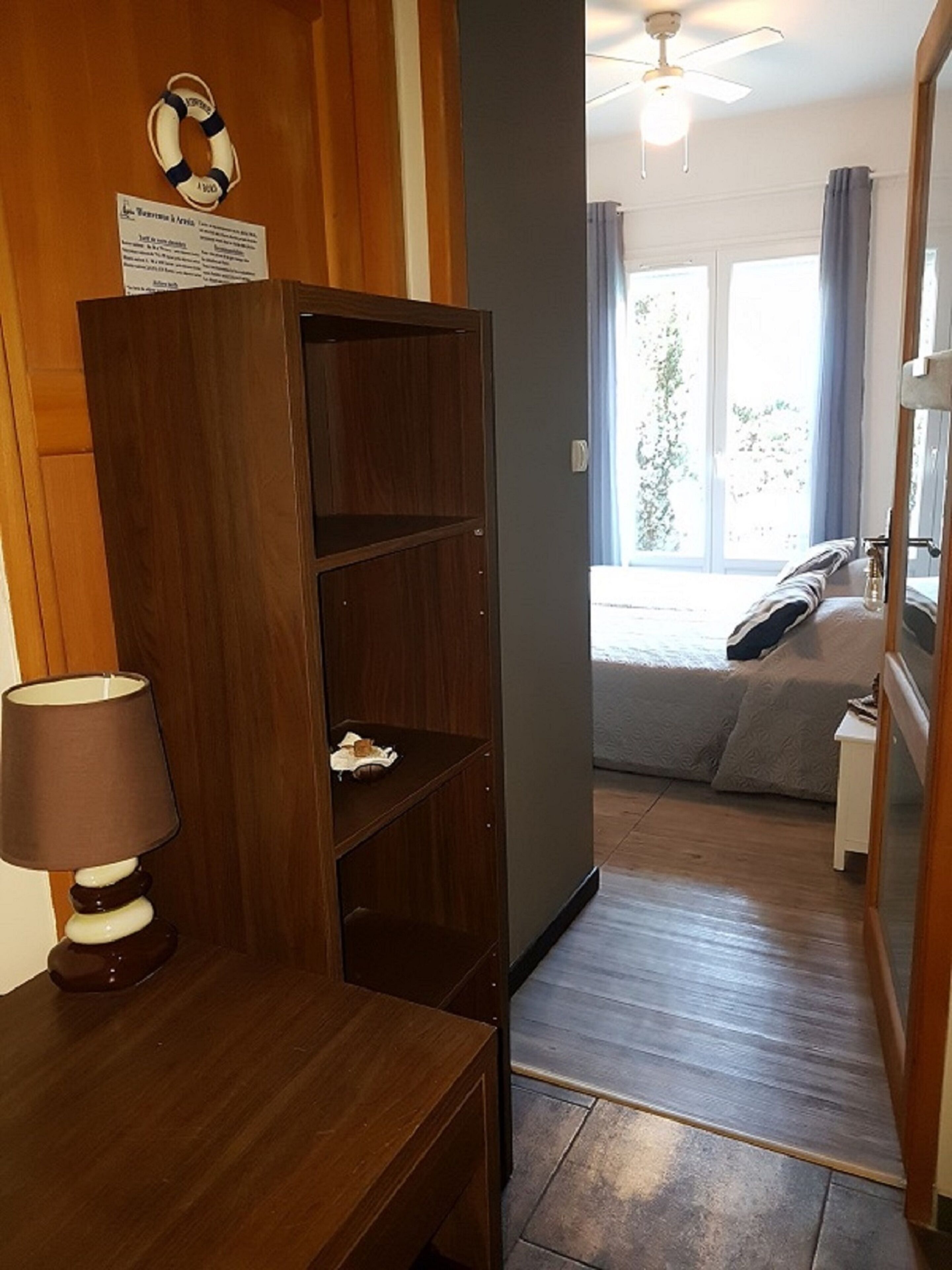 Quarto de hotel com escrivaninha de madeira, luminária, cama com vista para o exterior e ventilador de teto.