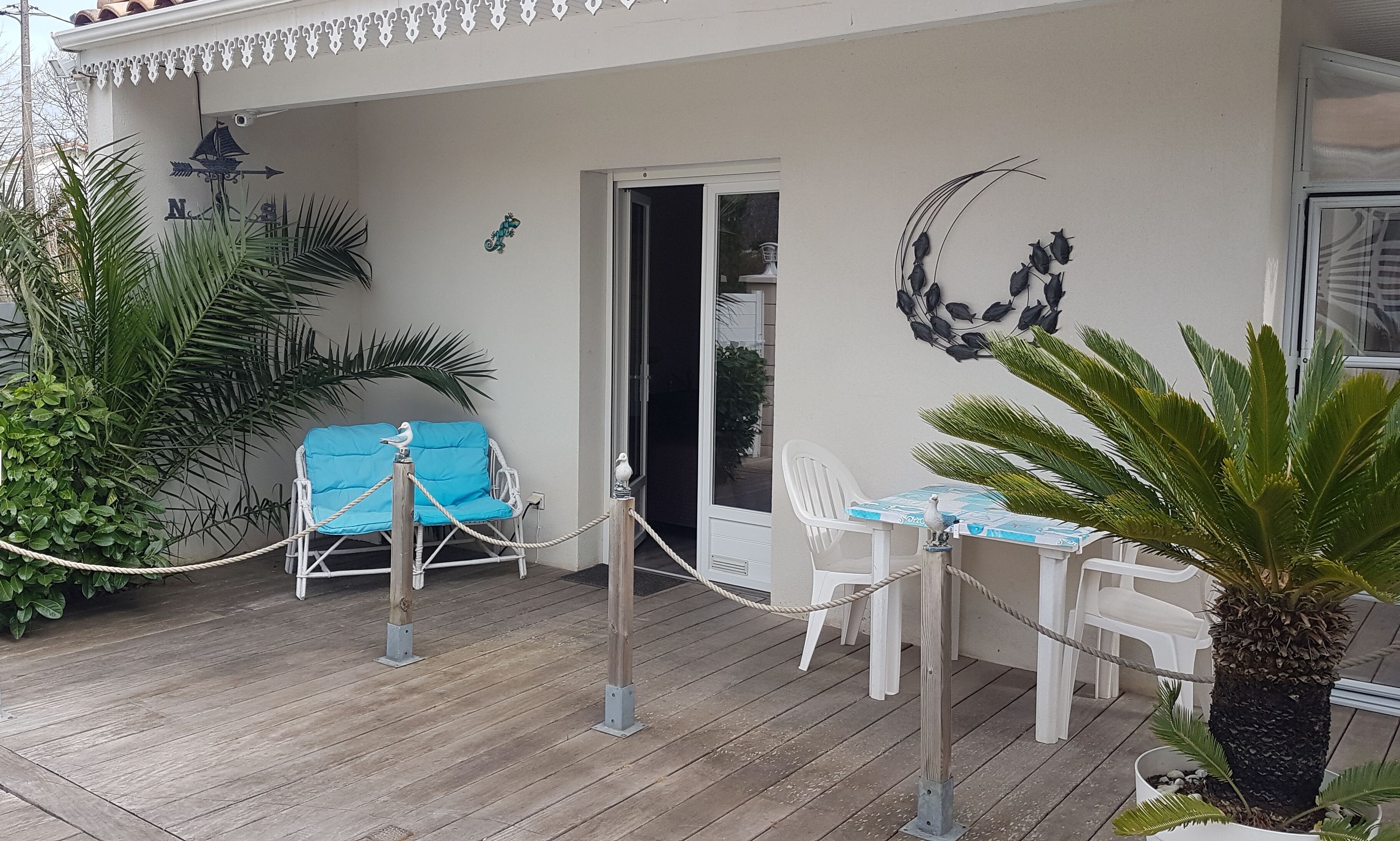 Un espace extérieur abrité doté d’une terrasse en bois, d’un fauteuil bleu rembourré, de chaises en plastique blanches et d’une plante en pot.