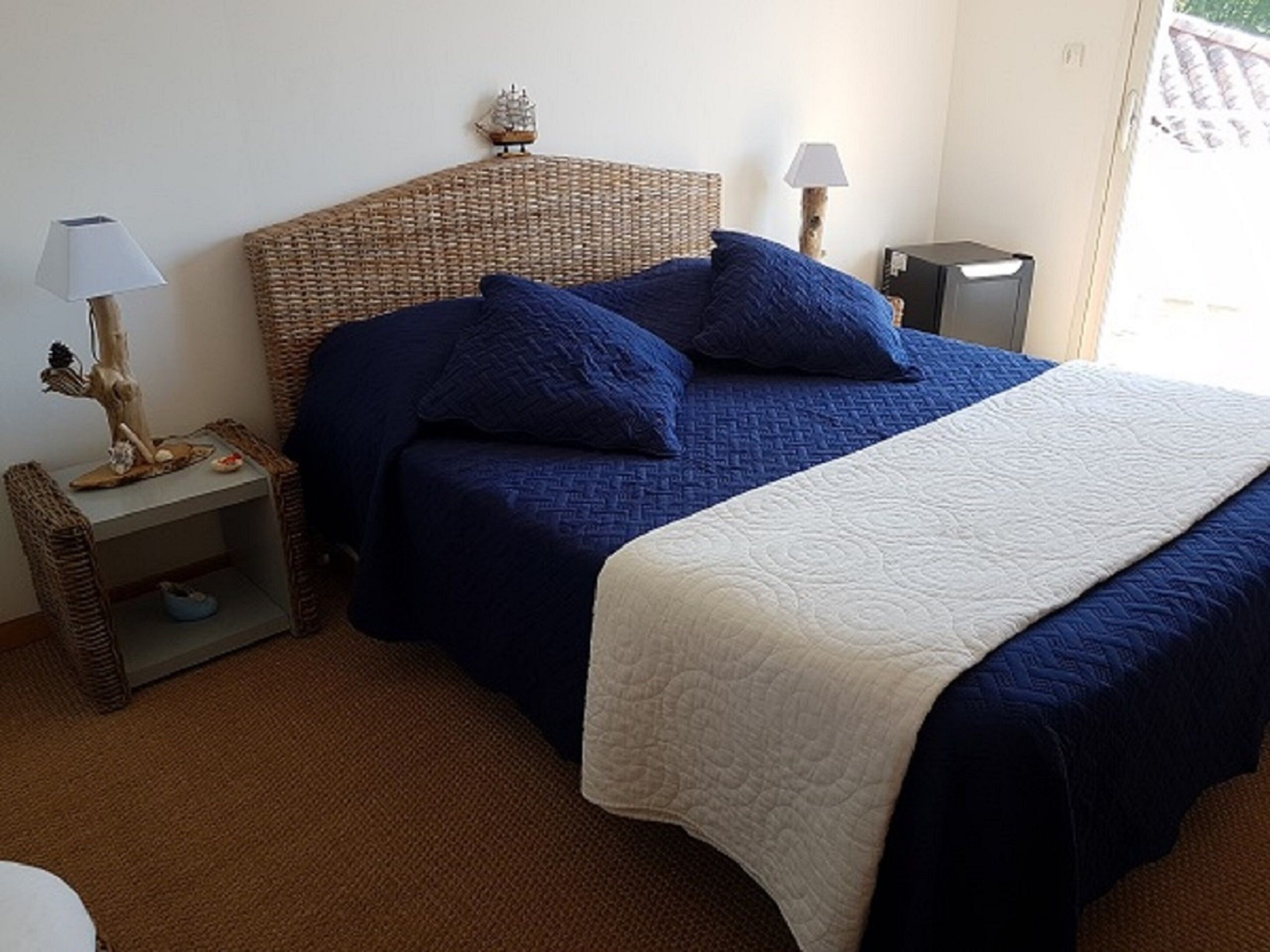 Quarto com cabeceira de vime, duas mesas de cabeceira com abajures, cama com roupa de cama azul e manta branca.