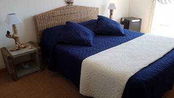 Un dormitorio con cabecera de mimbre, dos mesitas de noche con lámparas, cama con ropa de cama azul y una manta blanca.