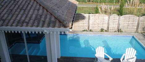A swimming pool with two white Adirondack chairs on a wooden deck.