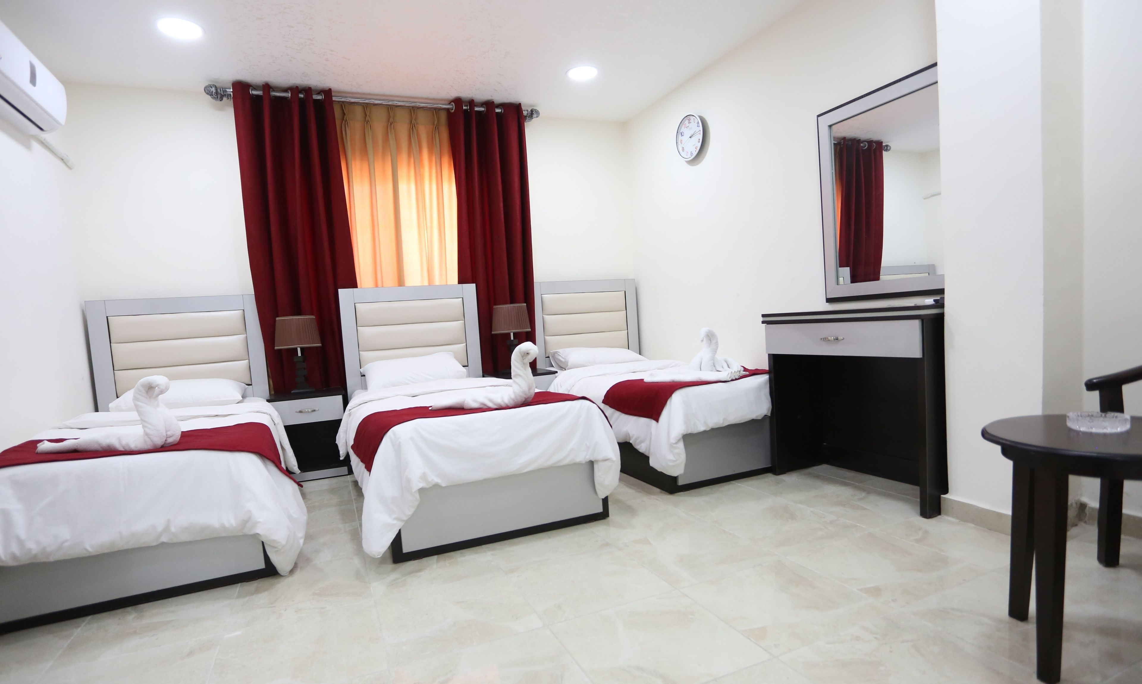 A hotel room with three beds, a desk, a mirror, and a clock on the wall.