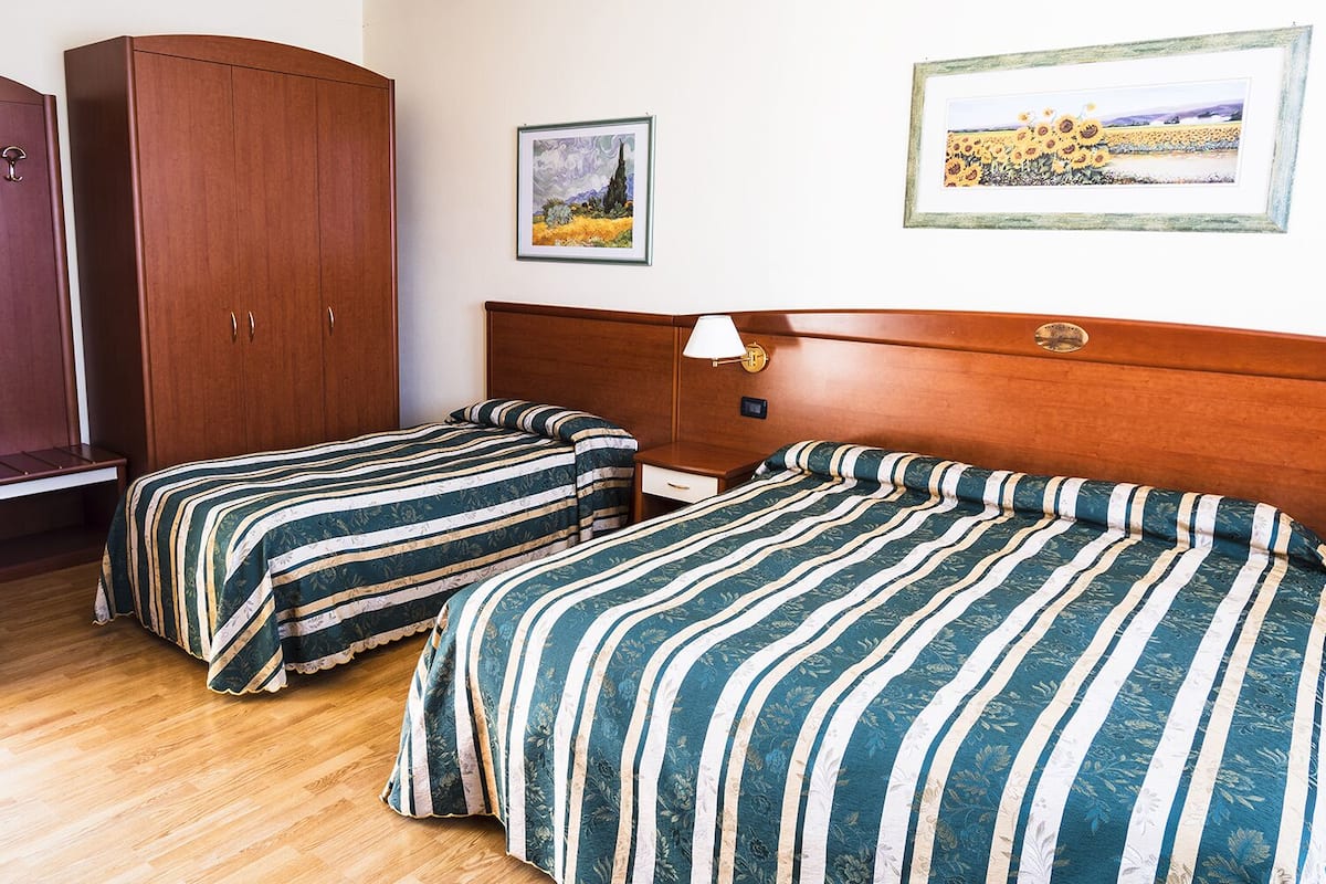 Une chambre d’hôtel avec deux lits simples, une armoire en bois, une table de chevet et deux œuvres d’art encadrées accrochées au mur.