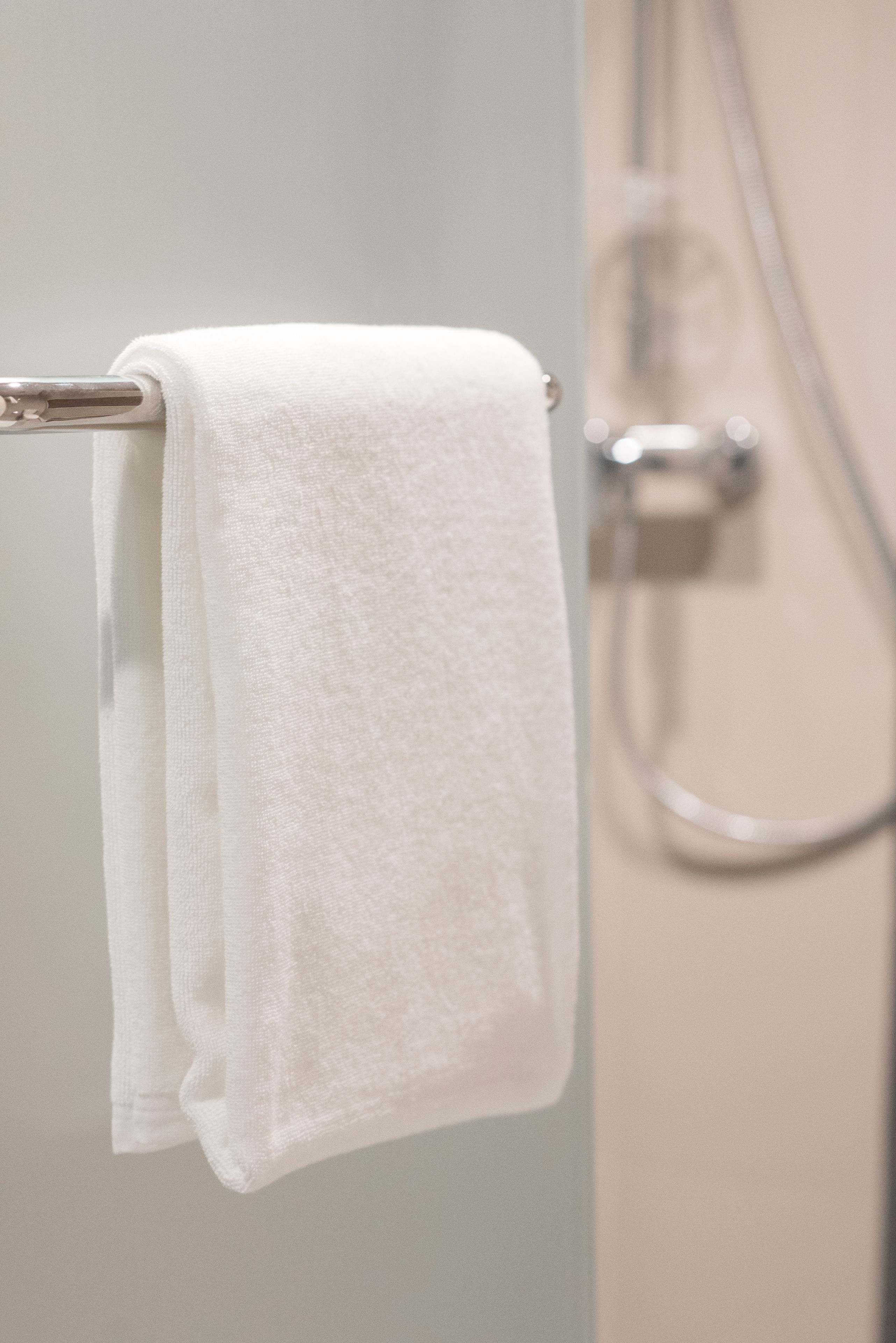 A white towel hanging on a metal towel rack in a bathroom with a visible shower head and control panel.